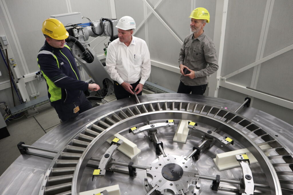 Eine in der Sonderlösung von Röhm gespannte Verdichter-Leitscheibe für Kraftwerksturbinen: Markus Zapke, Teamleiter im Bereich Equipment, Tooling und Processes bei Siemens Energy (r.), Matthias Barth, Sales Engineer und Fachberater bei Röhm (Mitte) mit Maschinenbediener Marvin Motza. Im Hintergrund ist die Roboteranlage zu sehen: Sie übernimmt die bisherige, körperlich anstrengende Tätigkeit. 