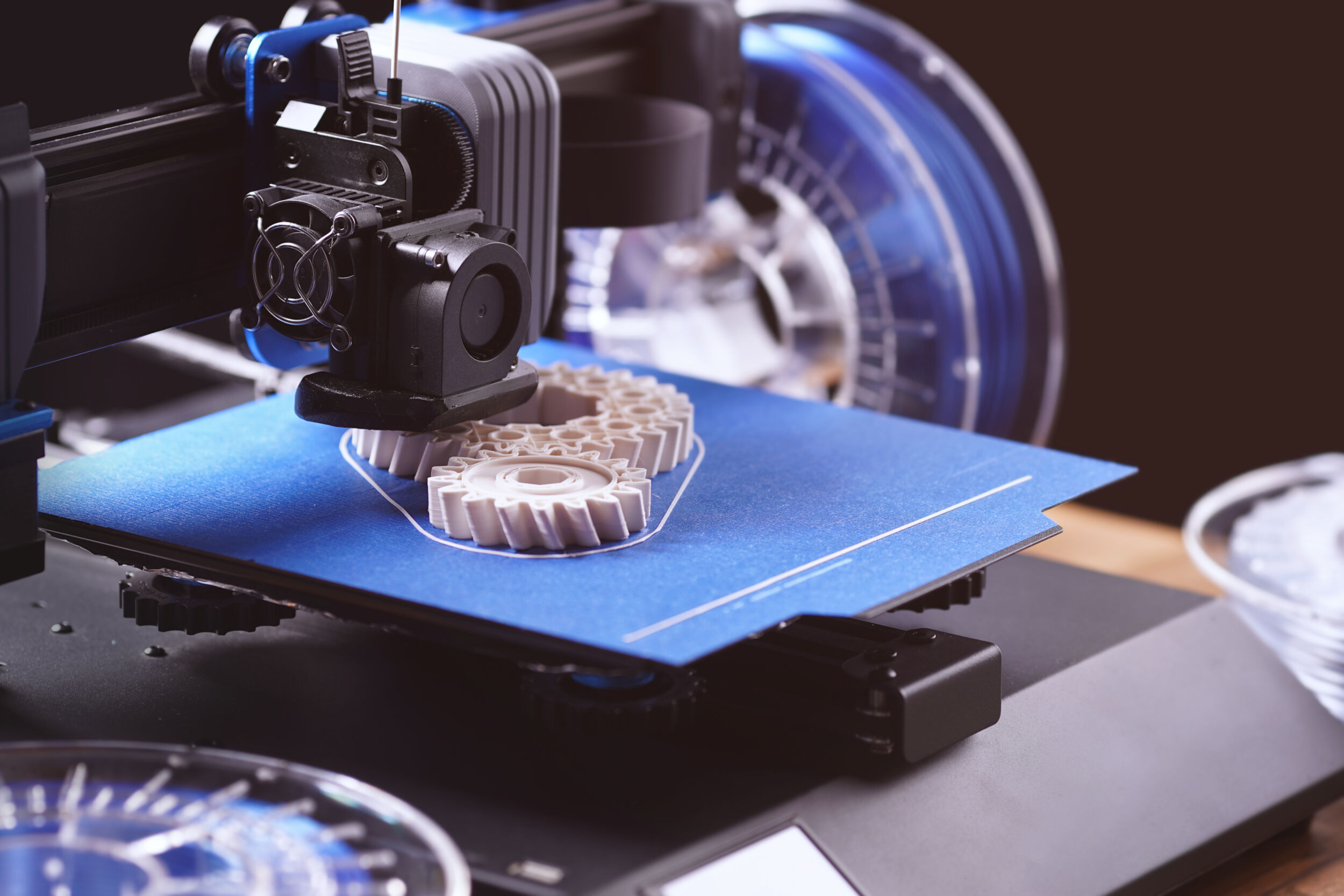 black FDM 3D printer on wooden table in bright warm light produces cream white helical gears. scenery in stylish dark surrounding with different filament rolls. additive manufacturing concept