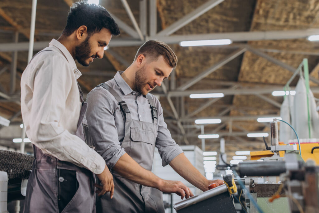 Mit gezielten Investitionen zusätzliches BIP pro Kopf generieren 1 A male inspector or operator of a workshop for the production of aluminum and plastic wreaths trains an intern. International team of men working together near a machine in a factory.