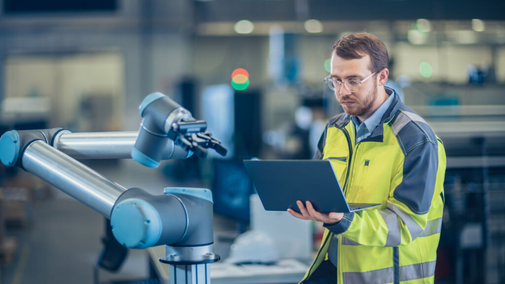 Hersteller investieren in neue Geschäftsmodelle 1 At the Factory: Automation Engineer Uses Laptop for Programming Robotic Arm. New Era in Automatic Manufacturing Industry.
