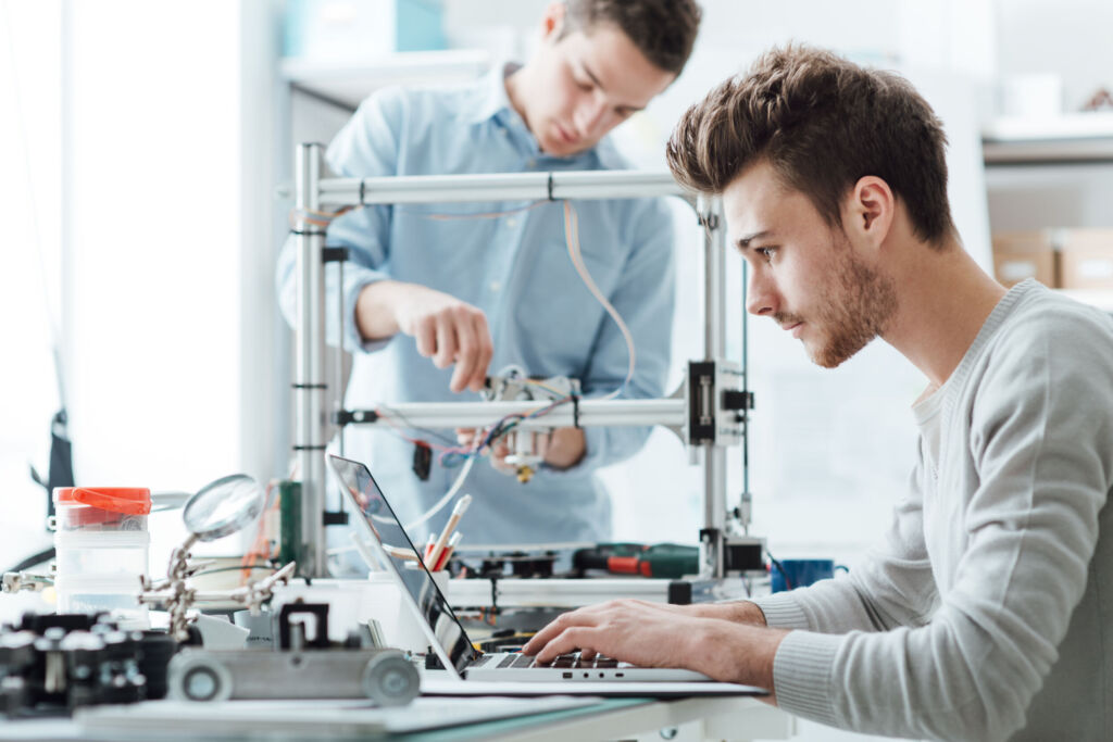 Deutsche Wirtschaft steigert Innovationsausgaben auf 190Mrd.€ 1 Engineering students working in the lab, a student is adjusting a 3D printer's components, the other one on foreground is using a laptop