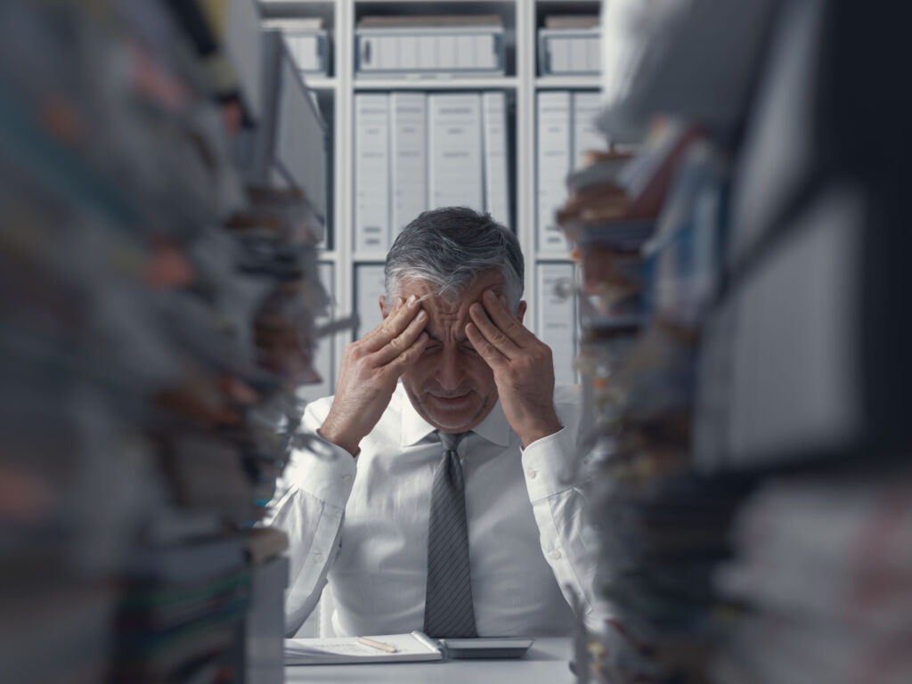 Innovationsschub und weniger Bürokratie 1 Stressed exhausted business executive in the office overloaded with work, he has piles of paperwork on the desk