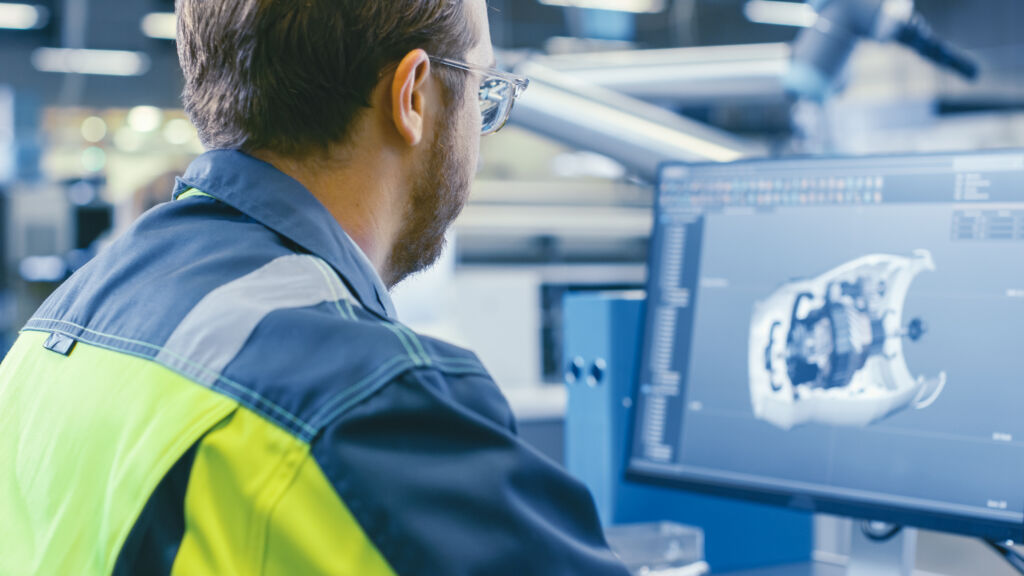 Der digitale Zwilling im Maschinen- und Anlagenbau 1 Back Shot of an Mechanical Engineer Working on Computers, Designing in 3D Model of the Engine using CAD Software. At the Factory.