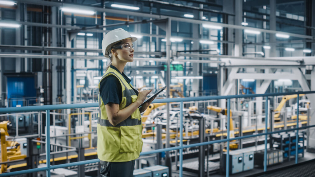 Wettbewerbsfähigkeit durch Modernisierung geschlossener Automatisierungssysteme steigern 1 Female Car Factory Engineer in High Visibility Vest Using Laptop Computer. Automotive Industrial Manufacturing Facility Working on Vehicle Production with Robotic Arms. Automated Assembly Plant