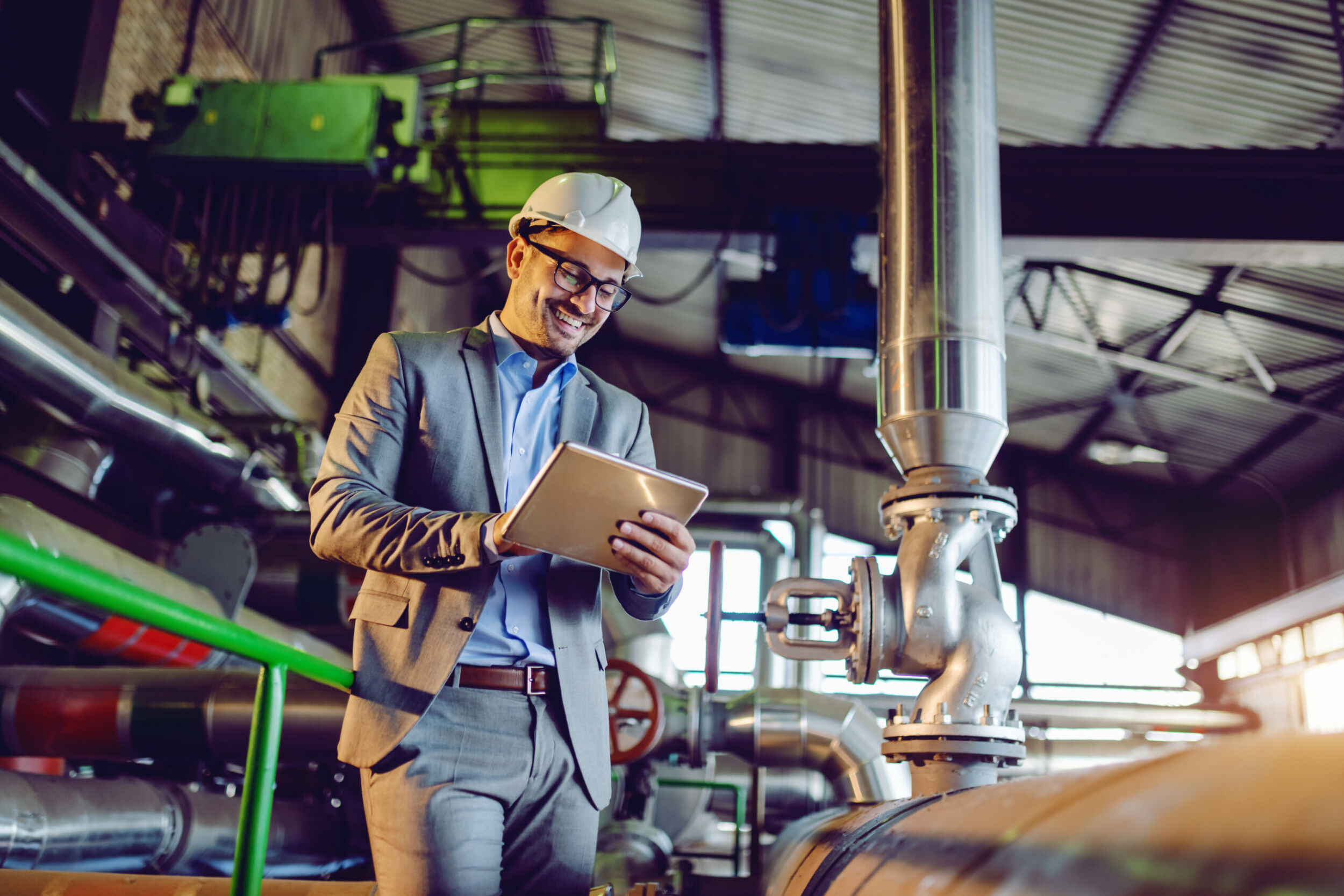 Handsome smiling caucasian supervisor in suit and with helmet on head using tablet while standing in power plant